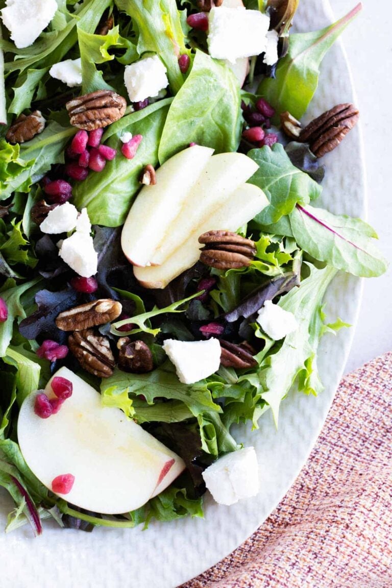 A fresh salad with mixed greens, apple slices, pecans, pomegranate seeds, and crumbled cheese on a white plate next to a woven napkin.