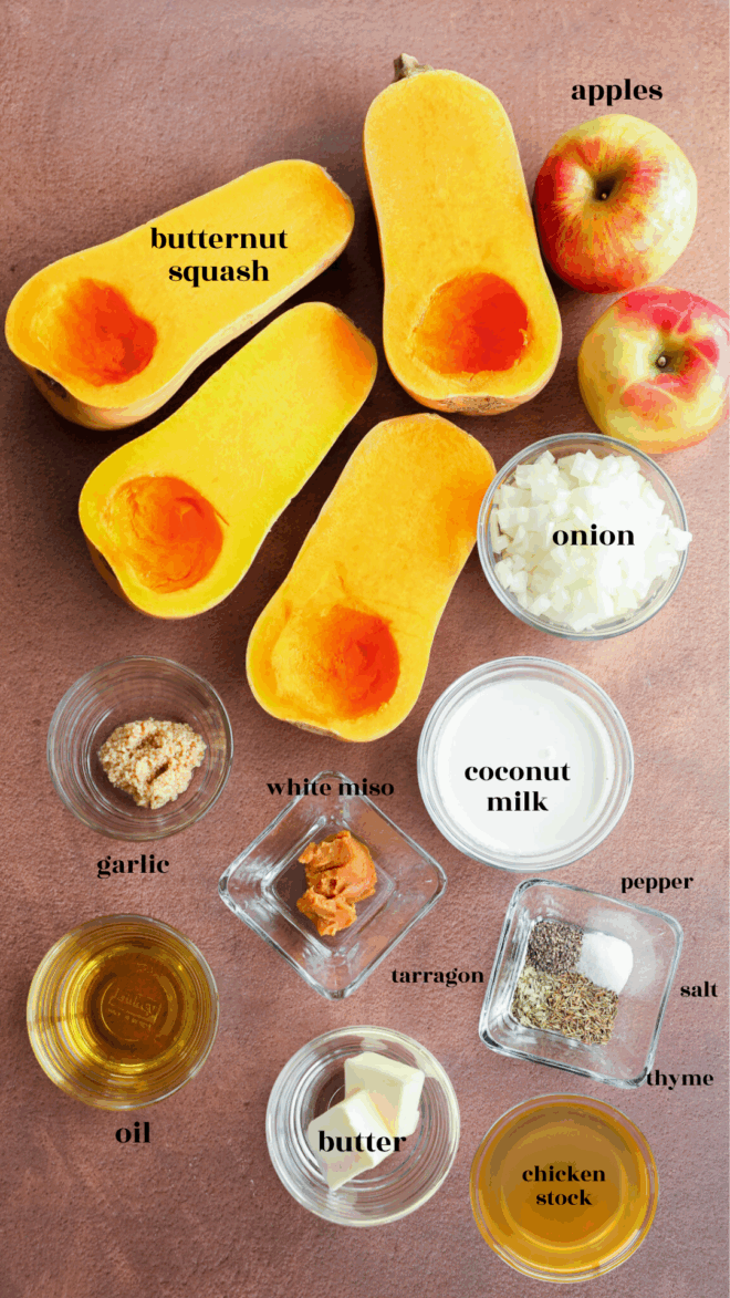 Ingredients for butternut squash and apple soup are arranged on a brown surface: halved butternut squash, apples, chopped onion, garlic, white miso, coconut milk, oil, butter, chicken stock, salt, pepper, tarragon, and thyme—all labeled.