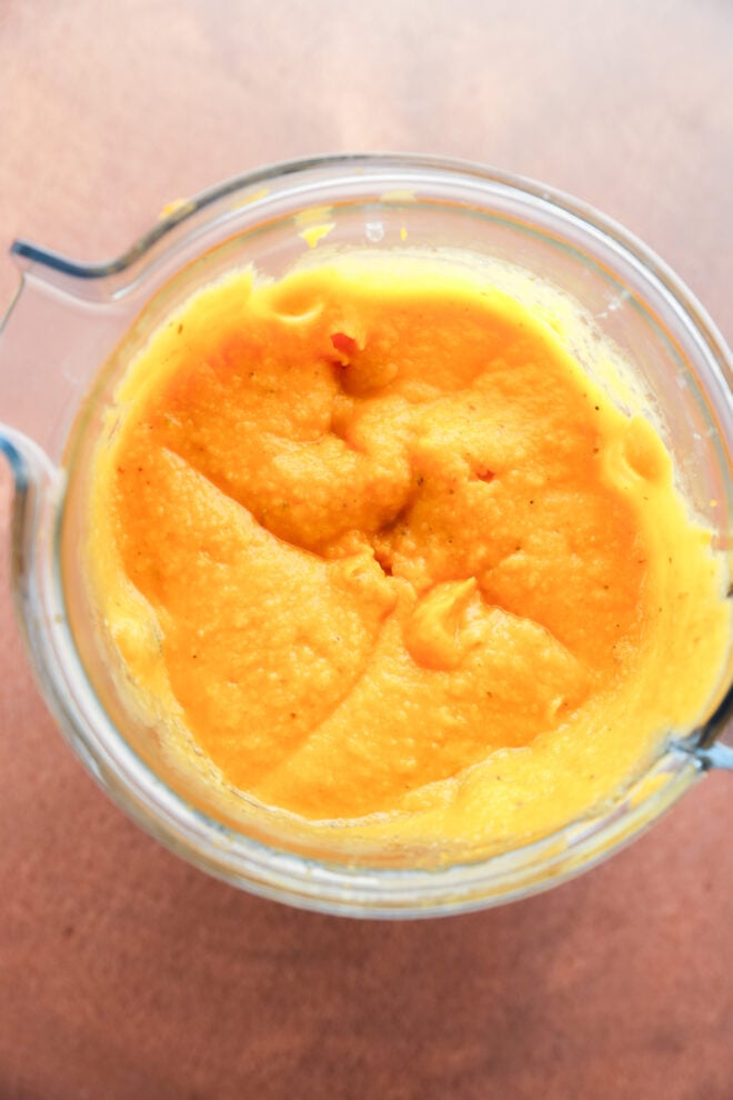 A blender filled with smooth, bright orange-yellow butternut squash puree sits on a light brown surface.