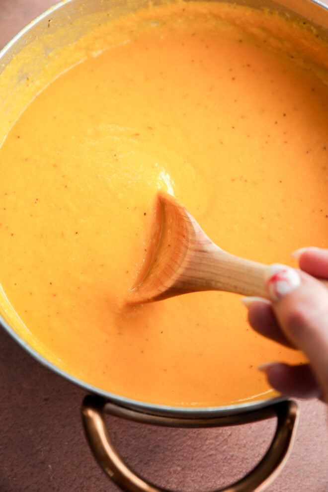 A hand stirs a thick, creamy butternut squash and apple soup in a metal pot using a wooden spoon. The soup appears smooth and rich.