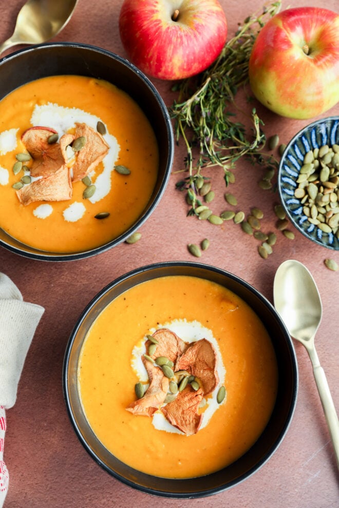 Two bowls of creamy fall soup, garnished with apple slices, pumpkin seeds, and cream, are surrounded by fresh apples, thyme, a bowl of pumpkin seeds, and two spoons on a brown surface.