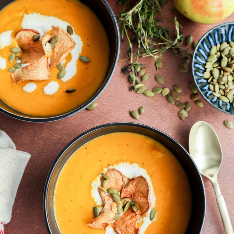 Two bowls of creamy fall soup, garnished with apple slices, pumpkin seeds, and cream, are surrounded by fresh apples, thyme, a bowl of pumpkin seeds, and two spoons on a brown surface.