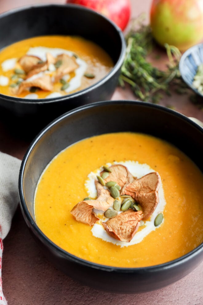 Two black bowls filled with creamy butternut squash and apple soup are topped with a swirl of cream, dried apple slices, and pumpkin seeds. Fresh herbs and whole apples are blurred in the background.