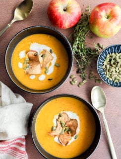 Two bowls of creamy butternut squash and apple soup, garnished with apple chips, pumpkin seeds, and a swirl of cream, sit on a brown surface with apples, fresh herbs, a dish of pumpkin seeds, and two spoons nearby.