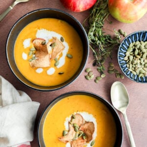 Two bowls of creamy butternut squash and apple soup, garnished with apple chips, pumpkin seeds, and a swirl of cream, sit on a brown surface with apples, fresh herbs, a dish of pumpkin seeds, and two spoons nearby.