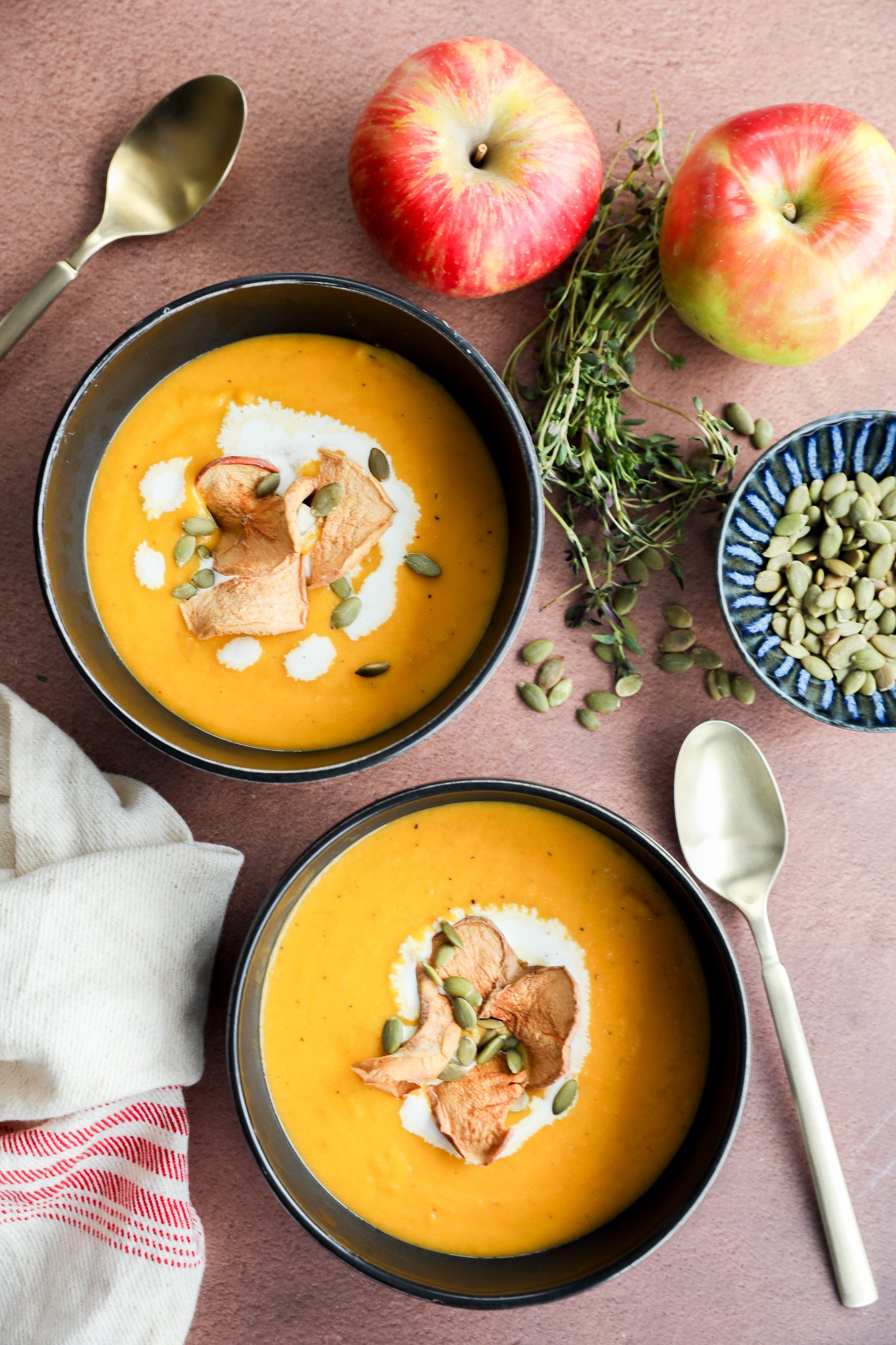 Two bowls of creamy butternut squash and apple soup, garnished with apple chips, pumpkin seeds, and a swirl of cream, sit on a brown surface with apples, fresh herbs, a dish of pumpkin seeds, and two spoons nearby.