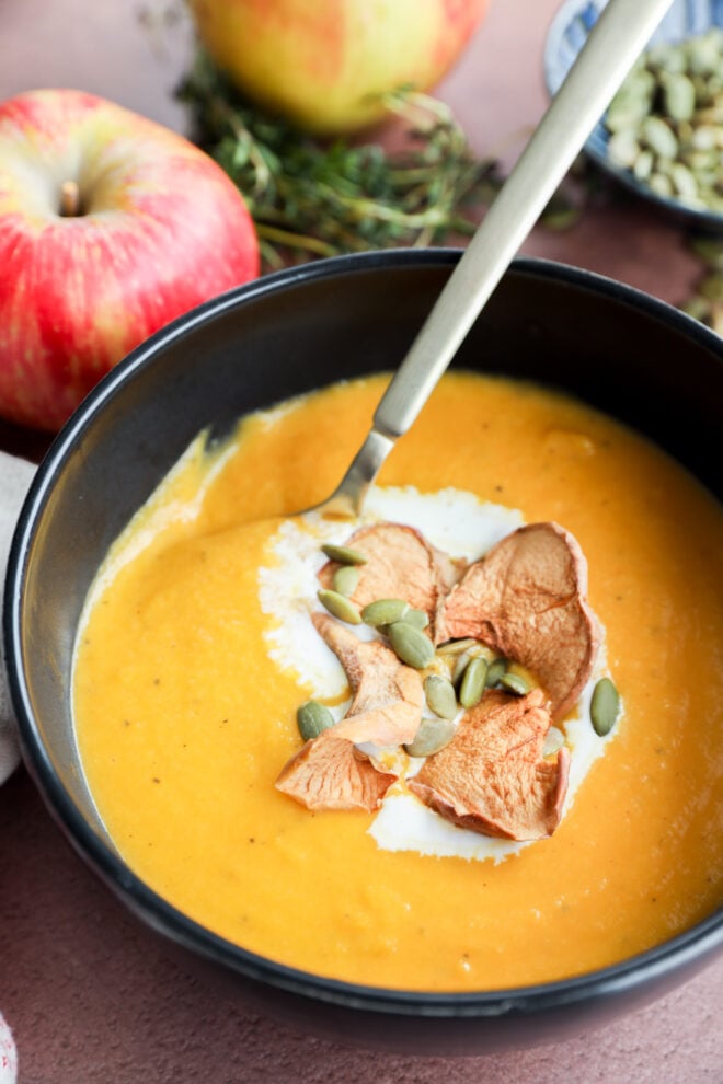 A bowl of creamy butternut squash and apple soup topped with dried apple slices and pumpkin seeds, with a spoon inside. An apple and herbs are in the background.