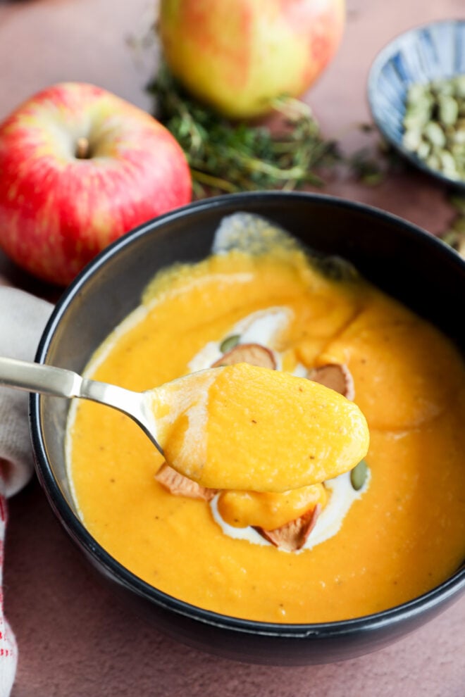 A spoonful of creamy butternut squash and apple soup is held above a black bowl filled with the same soup, garnished with seeds and cream. Fresh apples and herbs are in the background.