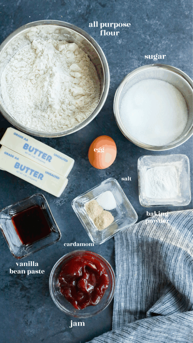 Top-down view of baking ingredients for christmas tree cookies on a dark surface: flour, sugar, two butter sticks, an egg, salt, baking powder, cardamom, vanilla bean paste, and a bowl of jam, with a striped kitchen towel beside them.