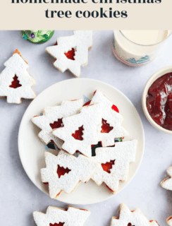 A plate of Christmas tree cookies dusted with powdered sugar, with a cutout tree design revealing red jam inside. A jar of jam, a mug, and festive decorations are in the background.