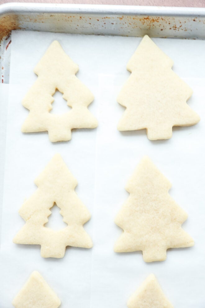 Christmas tree-shaped sugar cookies, some with small tree cutouts in the center, are arranged on a baking sheet lined with parchment paper.