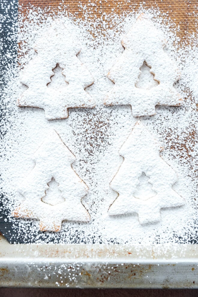 Four Christmas tree-shaped cookies are covered with a thick layer of powdered sugar on a baking sheet, with extra sugar sprinkled around them.