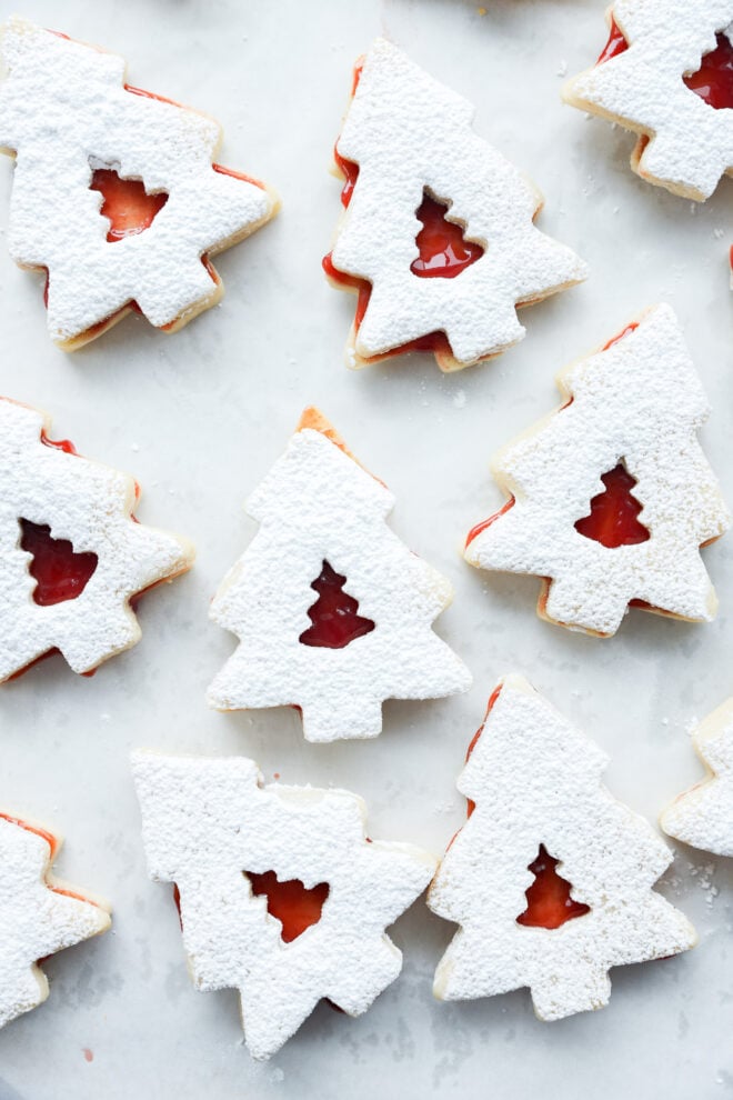 Christmas tree cookies dusted with powdered sugar, each with a small tree-shaped cutout in the center showing red jam filling underneath. The cookies are arranged on a light surface.