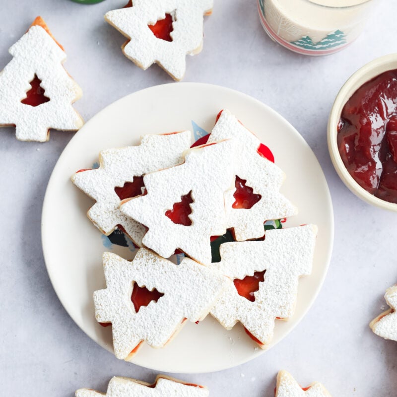 A plate of holiday cookies dusted with powdered sugar, filled with red jam, sits on a table. Nearby are more cookies, a glass of milk, a bowl of jam, and a small ceramic Christmas tree decoration.