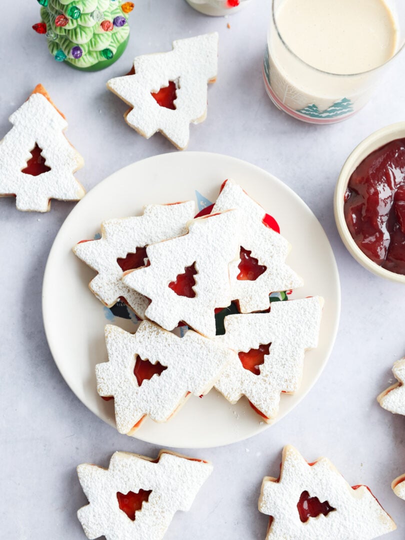 A plate of holiday cookies dusted with powdered sugar, filled with red jam, sits on a table. Nearby are more cookies, a glass of milk, a bowl of jam, and a small ceramic Christmas tree decoration.