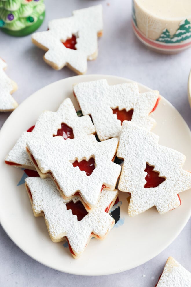 A plate of baked goodies dusted with powdered sugar, each with a cutout tree in the center revealing red jam filling, surrounded by festive decor.