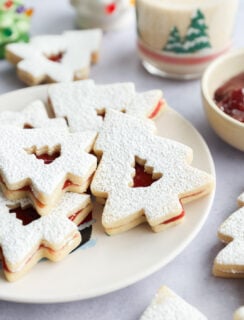 A plate of Christmas tree-shaped linzer cookies dusted with powdered sugar, filled with red jam. A glass with a festive tree design and a bowl of jam are in the background.