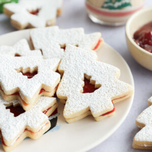 A plate of Christmas tree-shaped linzer cookies dusted with powdered sugar, filled with red jam. A glass with a festive tree design and a bowl of jam are in the background.