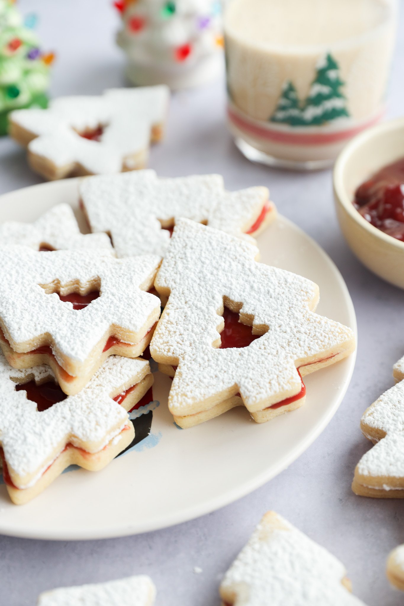 A plate of Christmas tree-shaped linzer cookies dusted with powdered sugar, filled with red jam. A glass with a festive tree design and a bowl of jam are in the background.