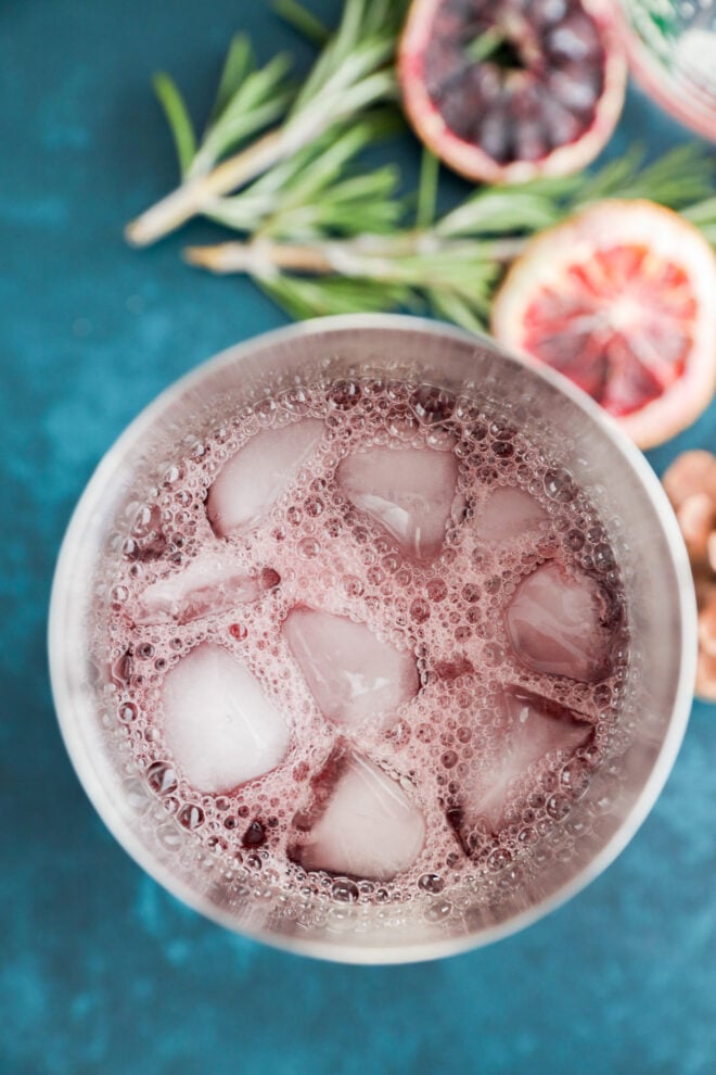 A close-up top view of a glass filled with a fizzy purple christmas vodka cocktail and ice cubes, set on a blue surface, with blood orange slices and sprigs of rosemary in the background.