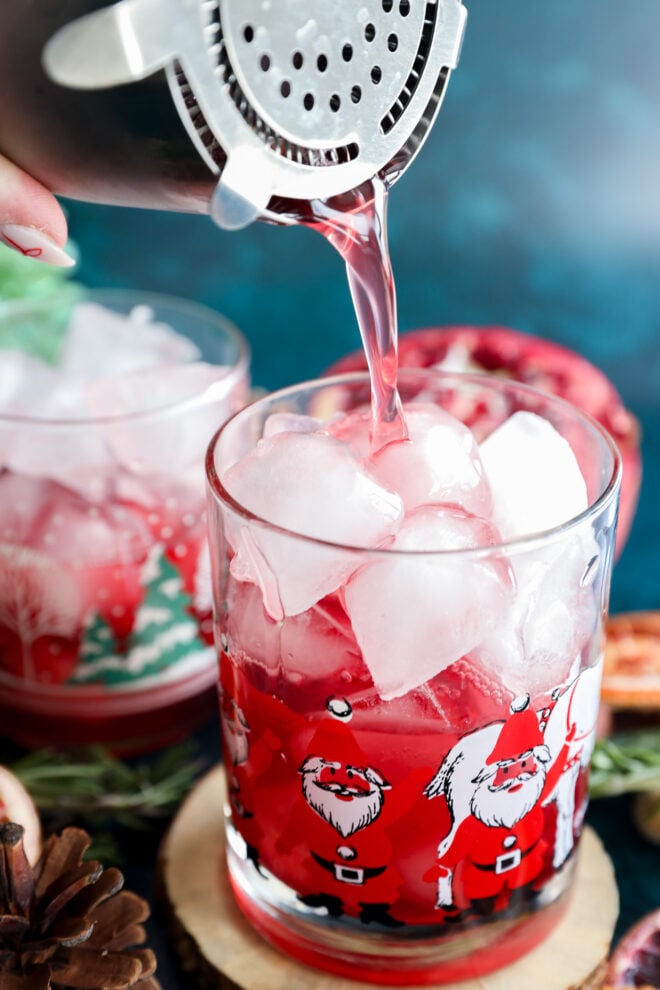 A close-up of a festive glass decorated with Santa Claus illustrations, filled with ice, as a pink-red christmas vodka cocktail is poured in using a shaker. Another similar glass and holiday décor appear in the background.
