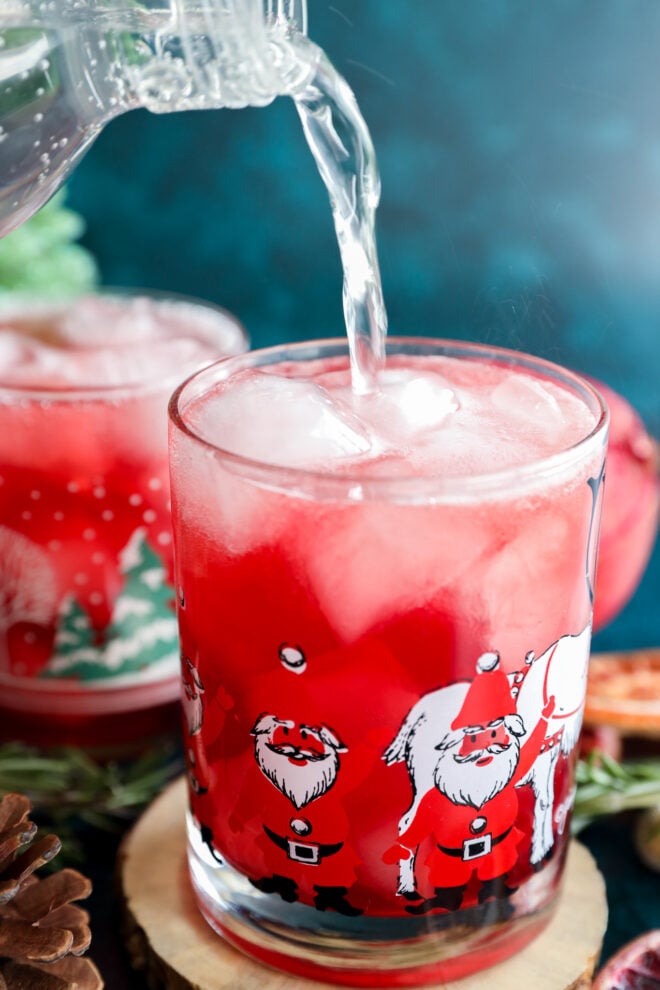 A clear liquid is being poured into a glass filled with ice and a bright red christmas vodka cocktail. The glass is decorated with images of Santa Claus, with another festive glass and decorations in the background.