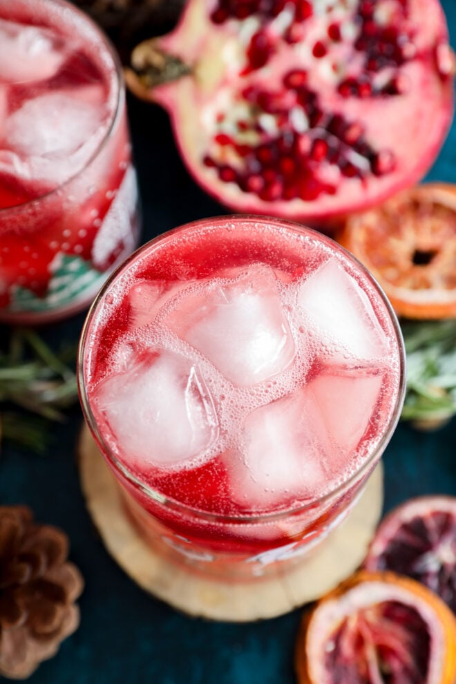 A close-up of a glass filled with a red, ice-filled christmas vodka cocktail, surrounded by pomegranate halves, dried orange slices, pinecones, and sprigs of rosemary on a dark background.