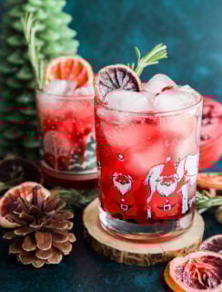A festive christmas vodka cocktail with ice, garnished with dried citrus and rosemary, served in a Santa Claus glass. Nearby sit a second similar drink, a pinecone, wooden coaster, dried fruit, and a green tree-shaped candle.