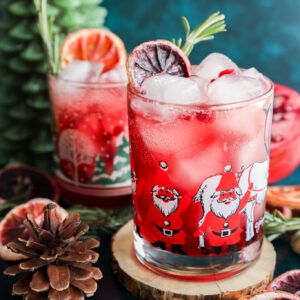 A festive christmas vodka cocktail with ice, garnished with dried citrus and rosemary, served in a Santa Claus glass. Nearby sit a second similar drink, a pinecone, wooden coaster, dried fruit, and a green tree-shaped candle.