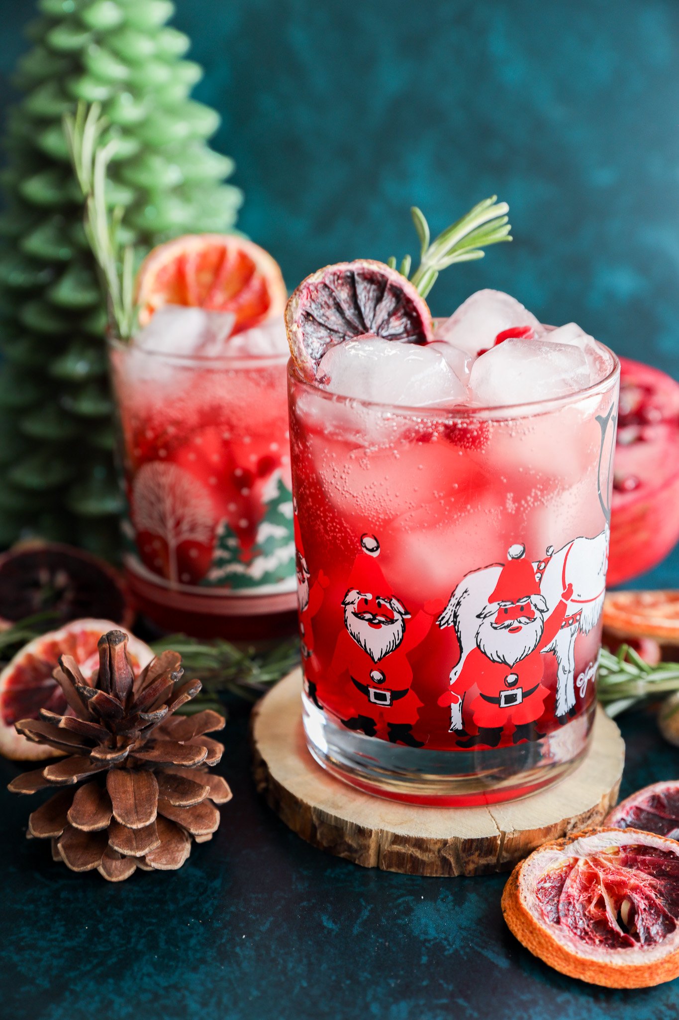 A festive christmas vodka cocktail with ice, garnished with dried citrus and rosemary, served in a Santa Claus glass. Nearby sit a second similar drink, a pinecone, wooden coaster, dried fruit, and a green tree-shaped candle.