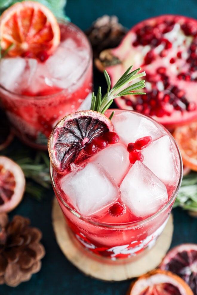 A glass of red christmas vodka cocktail with ice cubes, garnished with a slice of blood orange, pomegranate seeds, and a rosemary sprig, surrounded by another drink, dried citrus slices, and a halved pomegranate.