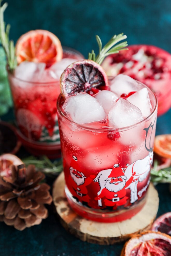 Two festive glasses filled with a Christmas vodka cocktail, ice cubes, and garnished with pomegranate seeds, rosemary, and dried citrus slices. The glasses feature Santa Claus illustrations, with pinecones nearby on a dark blue surface.