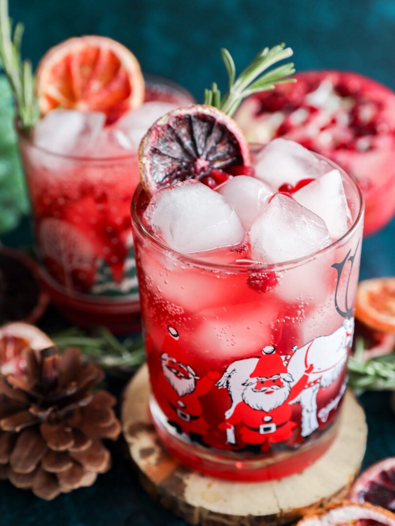 Two festive glasses filled with a Christmas vodka cocktail, ice cubes, and garnished with pomegranate seeds, rosemary, and dried citrus slices. The glasses feature Santa Claus illustrations, with pinecones nearby on a dark blue surface.