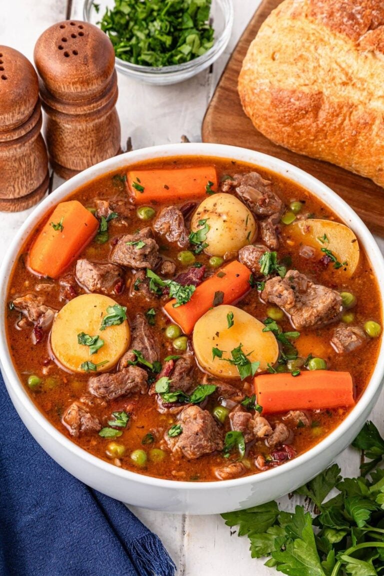 A bowl of beef stew with chunks of beef, potatoes, carrots, and peas in broth, garnished with parsley. Nearby are a loaf of bread, salt and pepper shakers, and a small bowl of chopped herbs.
