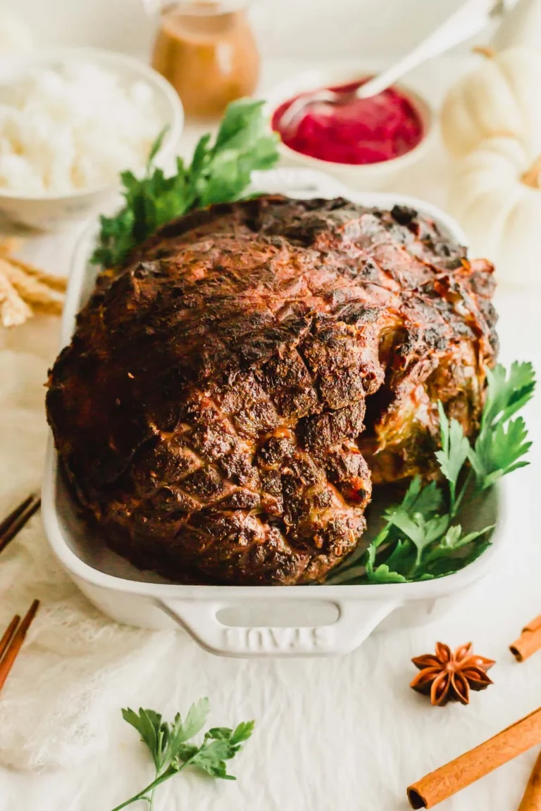 A large, roasted beef brisket sits in a white baking dish, garnished with fresh parsley. In the background, there are bowls of white rice, cranberry sauce, and gravy, with spices and herbs surrounding the dish.