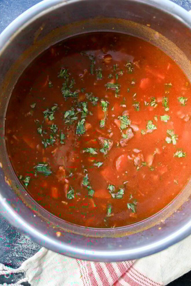 A pot of tomato-based soup or stew, reminiscent of Instant Pot Beef Bourguignon, with chopped herbs sprinkled on top, sits on a striped cloth.