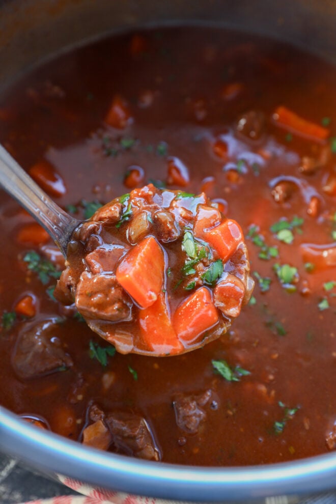 A close-up of a ladle lifting hearty Instant Pot Beef Bourguignon, with chunks of beef, carrots, and fresh herbs from a pot filled with rich, red broth.