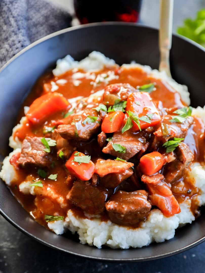 A bowl of mashed potatoes topped with Instant Pot Beef Bourguignon stew, featuring tender beef chunks, carrots, and rich brown gravy, garnished with chopped parsley. A spoon is visible in the bowl.