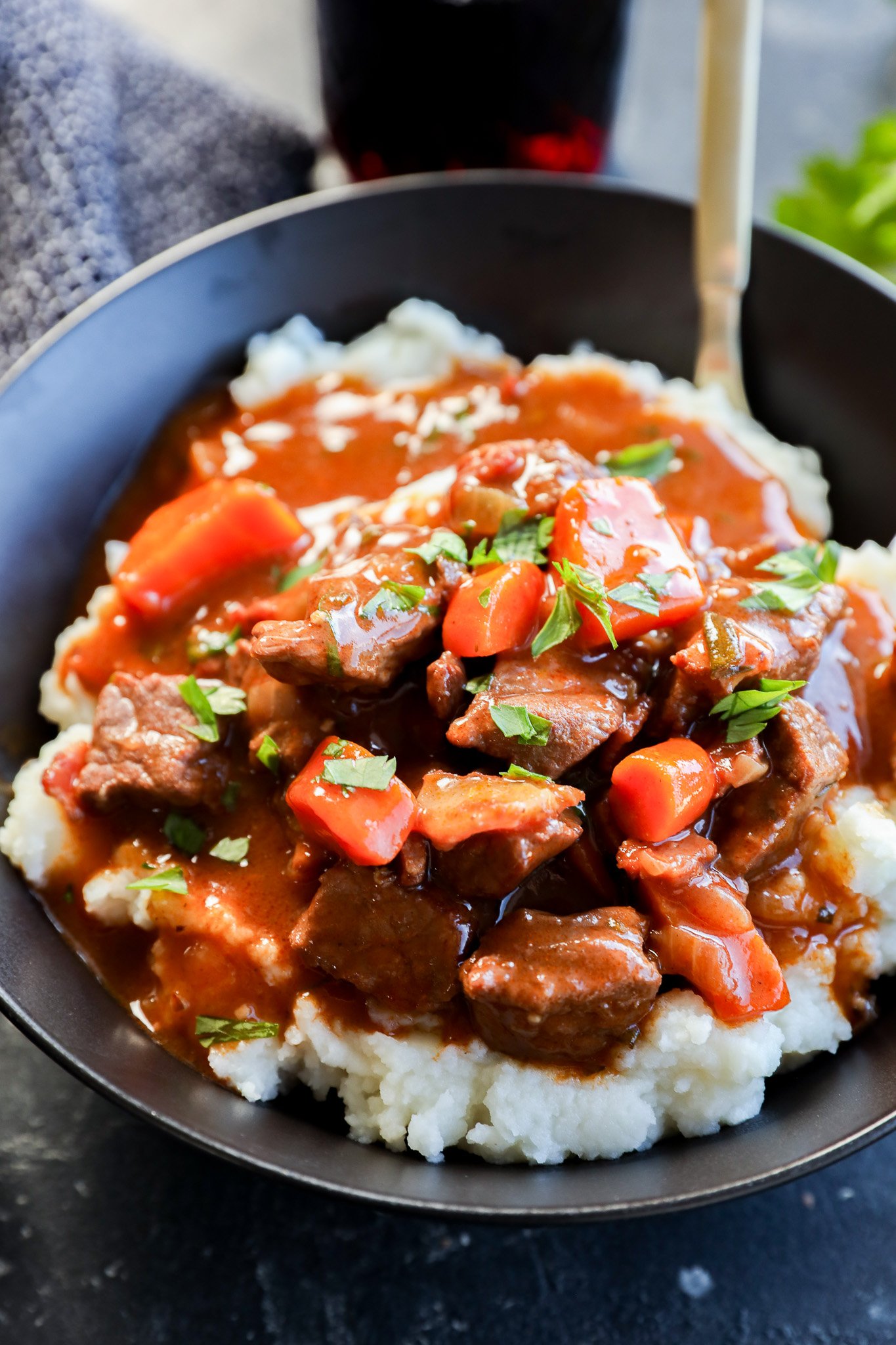 A bowl of mashed potatoes topped with Instant Pot Beef Bourguignon stew, featuring tender beef chunks, carrots, and rich brown gravy, garnished with chopped parsley. A spoon is visible in the bowl.