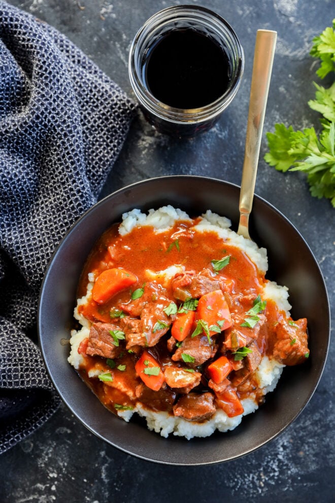 A bowl of mashed potatoes topped with savory Instant Pot Beef Bourguignon containing carrots and garnished with herbs, placed next to a glass of dark beverage, a gray cloth, and fresh parsley on a dark surface.