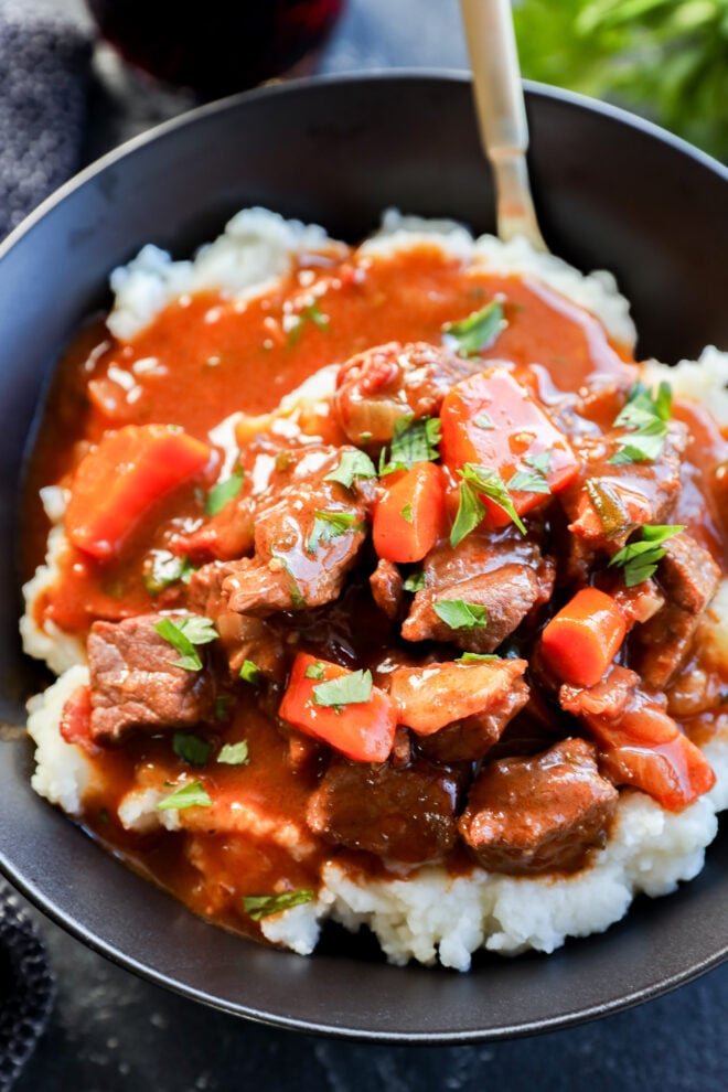 A black bowl filled with mashed potatoes topped with savory Instant Pot Beef Bourguignon, including chunks of beef, sliced carrots, and rich brown gravy, all garnished with chopped fresh herbs.