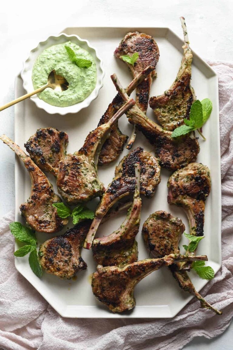 Grilled lamb chops arranged on a white platter, garnished with fresh mint leaves, served with a bowl of creamy green dipping sauce on the side.