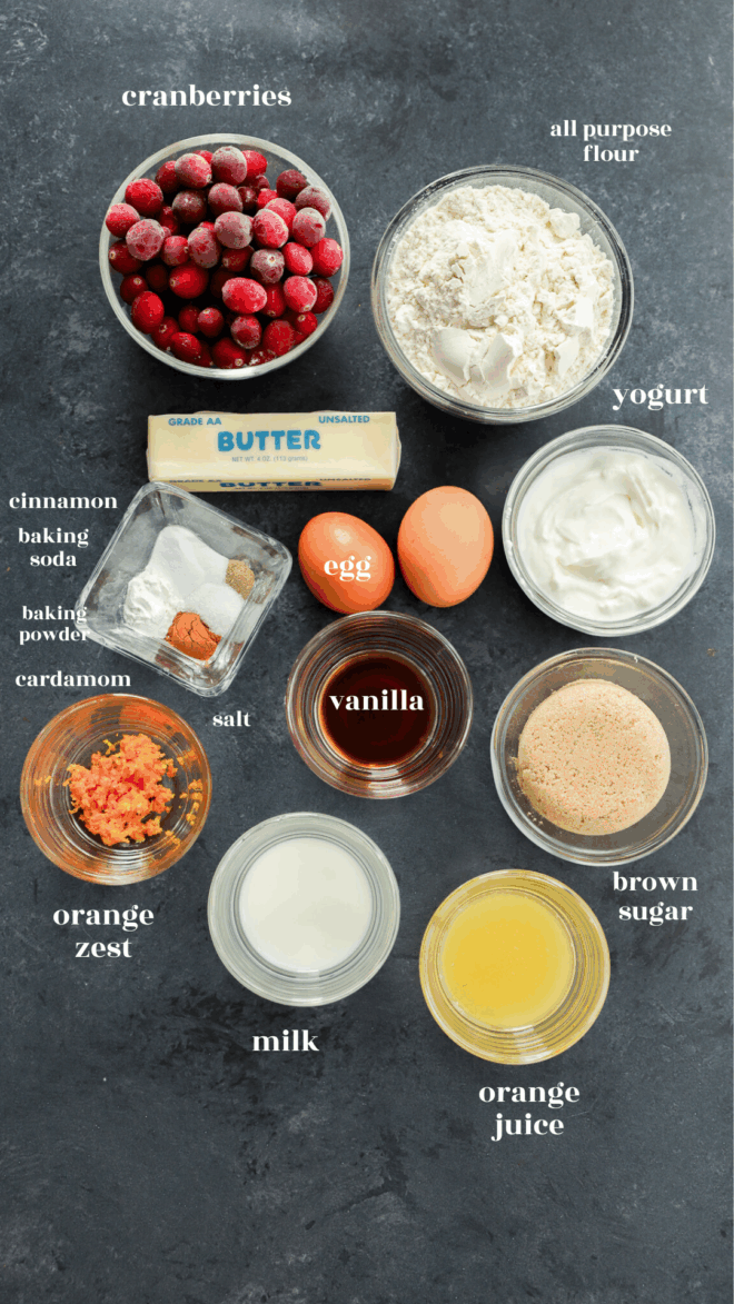 A flat lay of ingredients for orange cranberry muffins on a dark surface, including cranberries, flour, yogurt, butter, eggs, brown sugar, milk, orange juice and zest, vanilla, cinnamon, baking soda and powder, cardamom, and salt.
