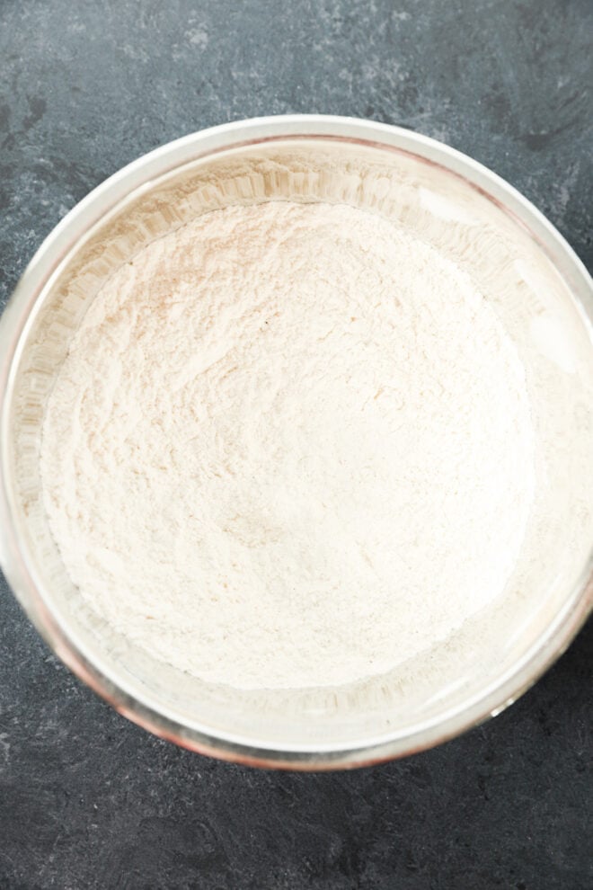 A glass bowl filled with white flour sits on a dark, textured surface, ready for baking.