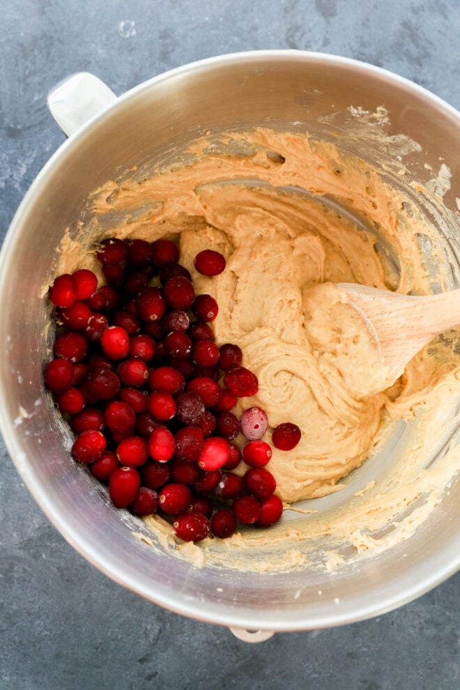 A metal mixing bowl containing thick batter and a pile of fresh red cranberries, with a wooden spoon resting inside, on a gray countertop.