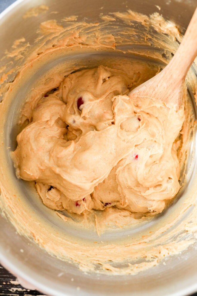A metal mixing bowl containing thick, creamy orange cranberry muffin batter being stirred with a wooden spoon.