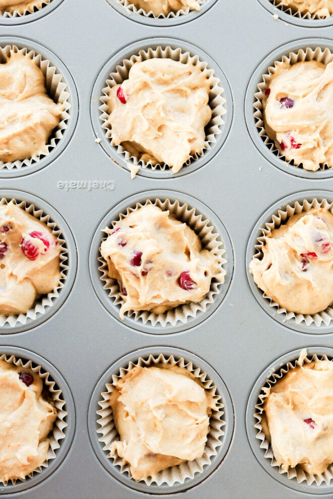 A muffin tin filled with paper liners holds unbaked batter, some with cranberries peeking through. The tin is viewed from above, and the batter is unevenly scooped into each cup.