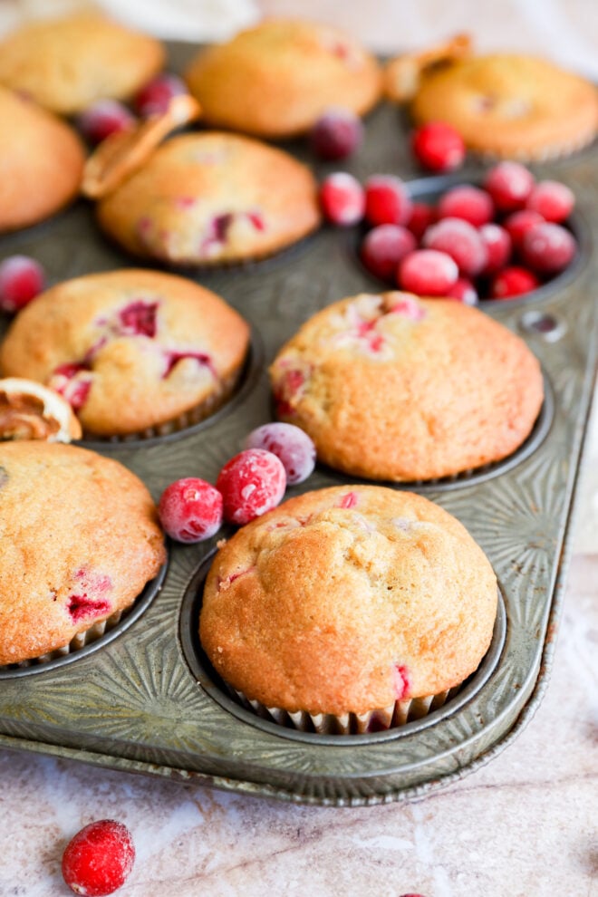 A close-up of golden-browned muffins in a muffin tin, with fresh cranberries scattered around. Some muffins have whole cranberries visible in the batter. The background shows a light, textured surface.
