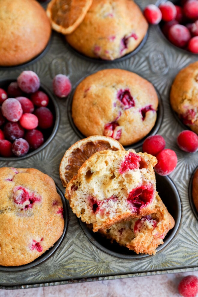 A muffin tin filled with golden brown orange cranberry muffins, some topped with dried orange slices and fresh cranberries, with one muffin broken open to reveal its moist interior and berry pieces.