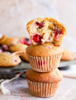 Two orange cranberry muffins are stacked on top of each other, with a third muffin—showing a bite taken out—resting on top. More muffins and scattered cranberries are in the background on a metal tray.